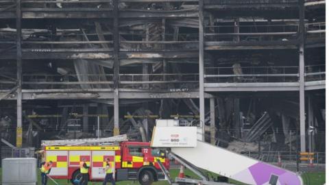 A wide shot shows the charred remains of cars in the soot covered car park.