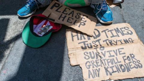 A homeless person begging for money with signs saying "please help" and "hard times, just trying to survive".
