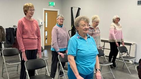 Elderly women standing up and being supported by chairs during an exercise class