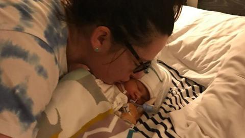 Sarah Robinson, who has dark brown hair pinned up and brown glasses, kisses her baby daughter Ida Lock on the cheek. The baby girl is wearing a white hat and is attached to tubes and wires.