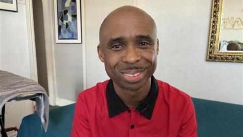 Ikenna Ajiero wearing a red shirt and sat on a sofa, smiling.
