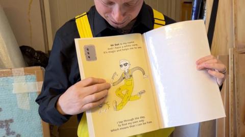 Mr Reynolds is in a different part of his studio and wears the same outfit. He is holding up an open section of his book and showing it to the camera. The page on the left side is a pale yellow and has an illustration of 'Mr Dot' who is wearing teh same outfit as Mr Reynolds and holding a white cane and walking. The text around the illustration says: "Mr Dot is my name and I have a white cane. It's magic and helps me to see. I tap through the day, to find my own way, which means that I can be free." The page on the right appears to be completely blank, but has the indents in the page for braille.
