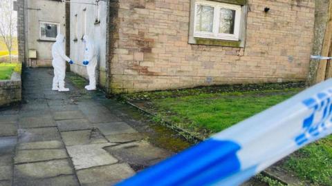 Two people in white forensic suits and blue gloves, outside a building, with blue and white police tape in the foreground.