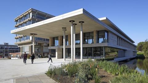 The Silberrad building at the University of Essex's Colchester campus. It is pictured on a sunny day with students walking in front of it. The building is cream and has columns in front of its many windows. In the background are other campus buildings