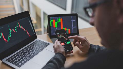 A man at home looking at charts on various different devices including a laptop, phone and tablet 