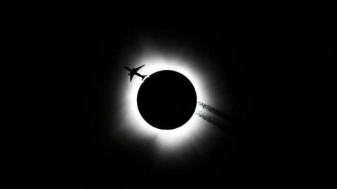 Black sky with white halo around a black disc. Silhouetted plane travelling through white halo