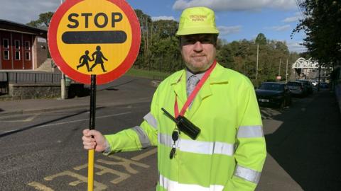 Tony Young outside Crumlin Integrated Primary School wearing shirt and tie and high vis jacket, with high vis hat that reads "crossing patrol" and lollipop stick sign with the word STOP on it