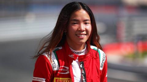 Chloe smiling, wearing red race overalls on a race track, with a white top. The overalls have advertising writing on the shoulders and across the front. The background is slightly blurred, with a streak of blue visible.