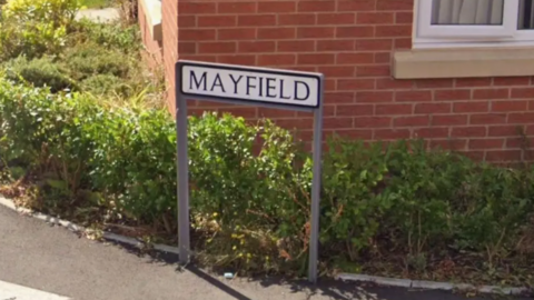 A road name of Mayfield can be seen in front of a small hedge and a red-bricked house.