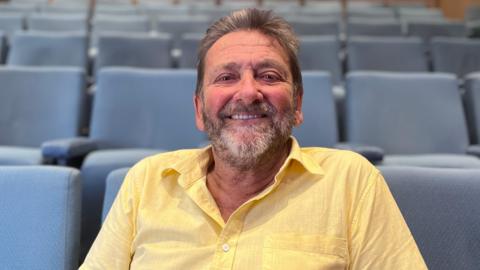 Gary is wearing a light yellow short-sleeved shirt sits among rows of blue auditorium-style seats. The image is taken in a lecture theatre or meeting room, with the seating and room interior softly out of focus behind Gary who is seated.