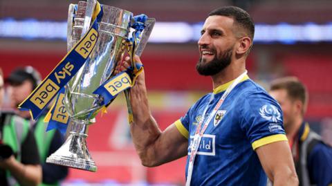 Omar Bugiel in blue AFC Wimbledon kit holds the League Two play-off final trophy, with its blue and yellow ribbons, aloft in his right hand