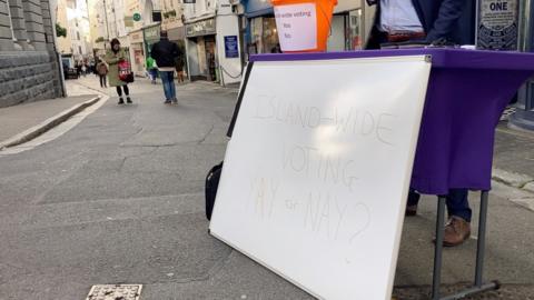 A white board which says Island-wide voting Yay or Nay? is resting on the front of a table with a purple tablecloth. It is outside in a road. There are pedestrians in the background.