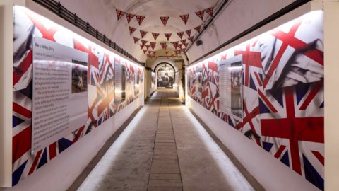 An exhibit inside Jersey War Tunnels. The displays have Union and Jersey flags dotted along them along with information boards. Union flag bunting is hanging from the tunnel's ceiling.