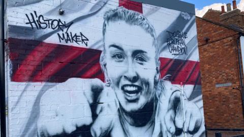 A large painting of Leah Williamson pointing with both hands is on the side of a brick wall. She is backed by the white and red England flat.