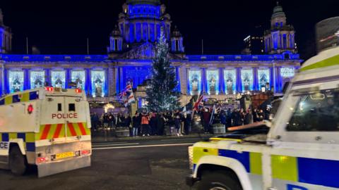 A blue lit Belfast City Hall. There is a Christmas tree in front and people carrying Union Jack flags have gathered at the gates. Two police land rovers are on the road.