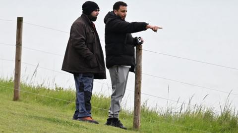 Walid Saadaoui (right) and Amar Hussein stand on a cliff edge, Saadaoui  leans against a wooden post in the ground that holds up a fence. He wears a black jacket, white t-shirt and grey jogging bottoms. Hussein wears a brown jacket and dark grey trousers