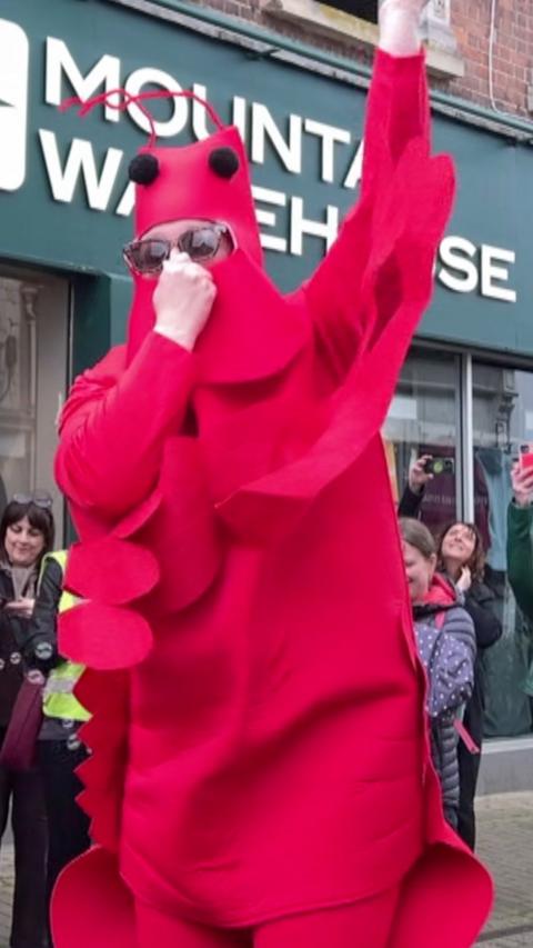 A man in a bright red lobster suit is holding his nose and has one arm in the air. He is standing in a high street.