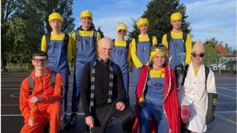 Nine students dressed in fancy dress, posing in two rows in a school playground.  Those standing at the back have yellow tops, hats and blue dungarees. Those in the front are kneeling. One wears an orange outfit and glasses, another wears black clothes, a bald cap and a long nose. A third wears a king's robes and crown. A fourth wears a white gown and glasses. 