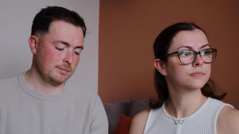 The McCreadys are sitting together on a sofa. Lauryn is looking wistfully out of a window while Andrew is looking downards towards their daughter's memory box
