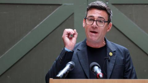 Andy Burnham, making a speech, wearing a dark T-shirt and grey jacket with a badge of a bee on the lapel. 
