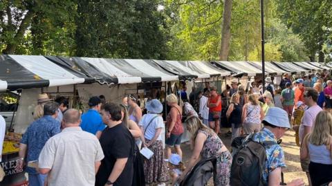 A crowd of people stood near black and white market stalls, with trees surrounding the area.