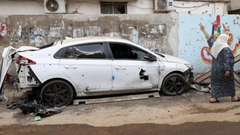 A woman walks past a damaged car in Jenin refugee camp in the occupied West Bank, following a deadly raid by Israeli forces (20 September 2023)
