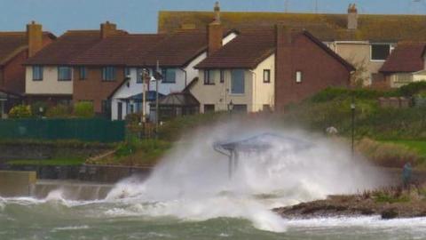 Waves and white foam shooting up onto the land in front of a cluster of houses