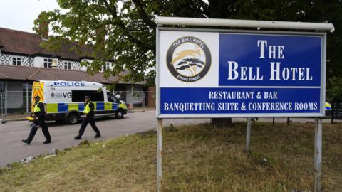 Two police officers walking next to the hotel, which is surrounded by a thin metal fence. A police van has also parked next to the building. In the foreground is a large blue sign which reads 'The Bell Hotel'. 