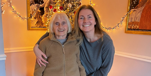 Two women standing close together, the younger woman on the right with her arm around the shorter older woman to our left. The woman on right is wearing a grey top and has brown shoulder length hair, while her mum has a beige zip-up fleece and grey hair in a bob. 