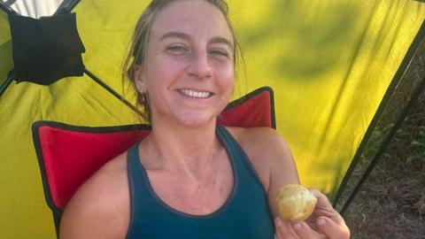 Emily Torode, sitting on a red chair and smiling in a green vest top, is seen holding a potato
