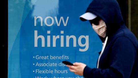 man wearing a face mask walks past a sign "Now Hiring" in front of a store amid the coronavirus pandemic on May 14, 2020 in Arlington, Virginia.