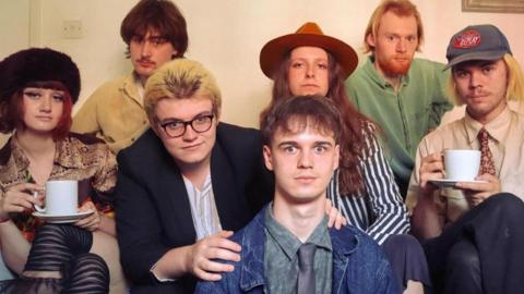 Magnolia the band pose for the camera - made up of seven male and female members sitting down - with two of the members at either end of the photo holding cups and saucers. 