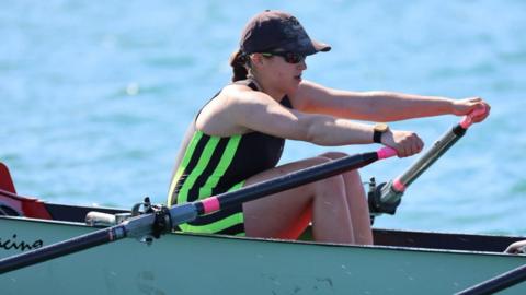 Ryanna is wearing a cap and sunglasses rows a racing boat on calm blue water, pulling an oar with pink grips, while another rower is partially visible alongside in matching striped kit.