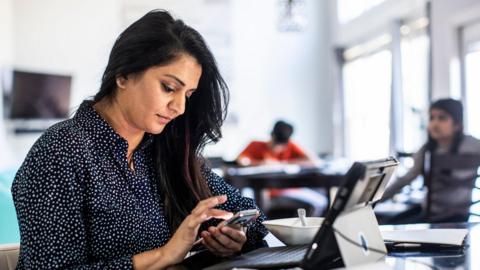 A female office worker sitting in front of a laptop and looking at her phone