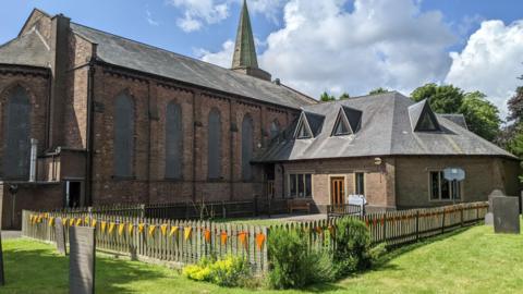 A brown brick church building with large windows and a spire in the background. In front of the church is a fence with bunting hanging from it, and some grass with gravestones on it