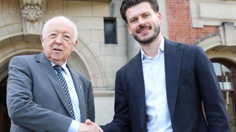 Two men in formal suits shaking hands outside a large stone building with arched doorways.
