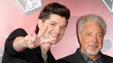 Danny O'Donoghue (left) and Sir Tom Jones (right) standing next to each other. They are both looking at the camera. Danny is doing a peace sign pose. They are both smiling.