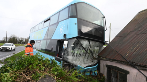 Blue double decker bus with smashed windscreen, crashed against a white property, large tree lying in front of it and a man in orange hi viz examining the scene 