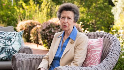 Her Royal Highness is sitting outside in a rattan garden seat. She is wearing a cream blazer with a blue shirt. She is also wearing a gold brooch. It is a sunny day with lots of greenery in the garden behind.