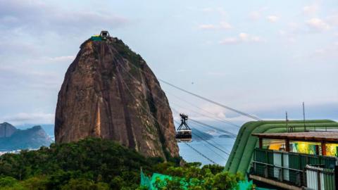 A view of Sugar Loaf cable car at Guanabara Bay in November 2024. The steep rocky mountain can be seen against the blue of the bay. At the top of the mountain, the cable car station is visible. Cables connect it with Morro da Urca, from which the photo is taken. A cable car can be seen suspended halfway between the two. 