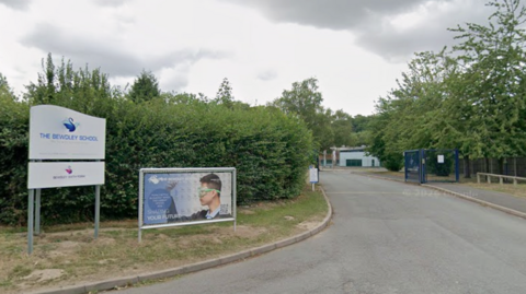 The entrance to the school which is a long driveway surrounded by trees and bushes on each side. There are dark blue metal gates open and buildings in the distance. On some grass to the left of the entrance are two signs. One is white with a swan logo and reads "the bewdley school" and the other has a young man with scientific googles on, looking at some equipment.