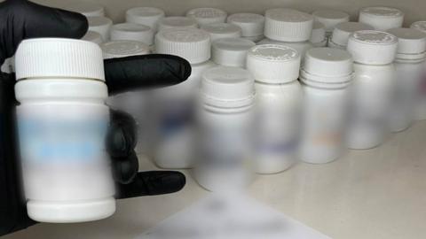 Black glove holding bottle of pills with more bottles in the background