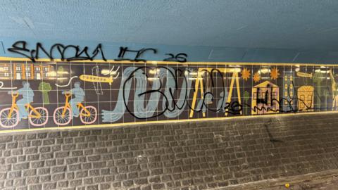 Art on the wall of an underpass, which depicts colourful cartoon figures riding bikes and playing on swings against a black background. However, a large graffiti tag has been sprayed over the top.