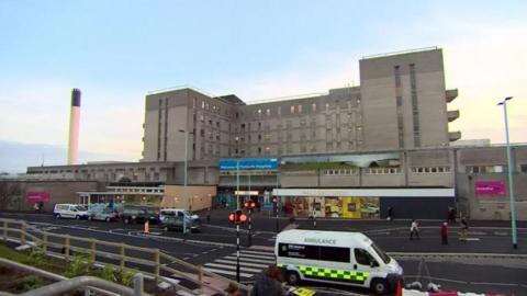 Derriford Hospital. It is a large concrete building with a road running in front of it which has an ambulance driving along it.