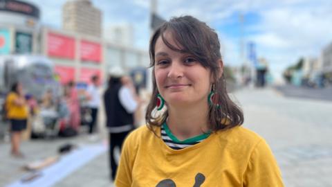 Cat a mother of two wearing a yellow t shirt with brown shoulder length hair and water melon earrings, behind her is a crowd with placards outside Churchill Square shopping centre