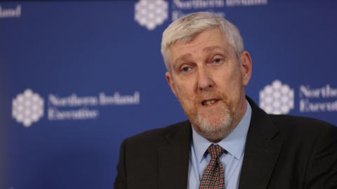 Finance Minister John O'Dowd, a man with short grey hair, wearing a black suit, blue shirt and red, yellow and navy tie. 