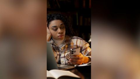 A woman checks her phone in the library. She is wearing a checkered shirt.
