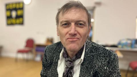 A man wearing a shirt and cravat-like tie sits in a room, looking at the camera. Chairs and posters can be seen in the background.