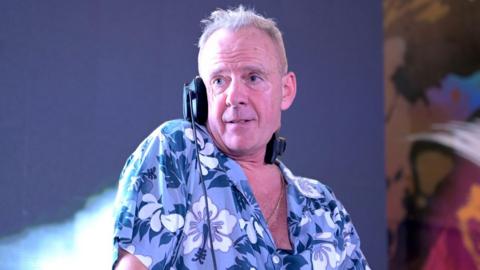 Fatboy Slim, Norman Cook, stands on stage with one ear of his overhead headphones on. He is looking off to the side. He is an older man with grey, short hair and is wearing a light blue, dark blue and white hawaiian shirt.