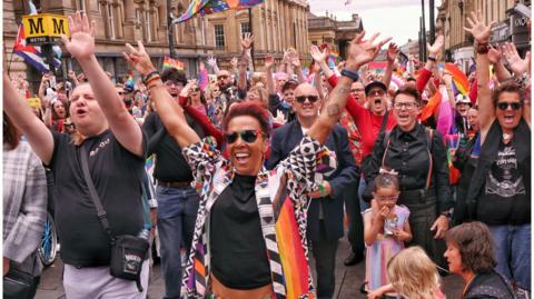 Kelly Holmes standing in a crowd of people on Grey Street, most of who have their hands in the air. She is smiling with her mouth open and is wearing a geometric jacket with the rainbow flag on it. She has short, dyed red hair and is wearing sunglasses. Everyone around her looks to cheering with their mouths open. There are rainbow flags throughout the crowd and the yellow Metro sign can be seen on the left.